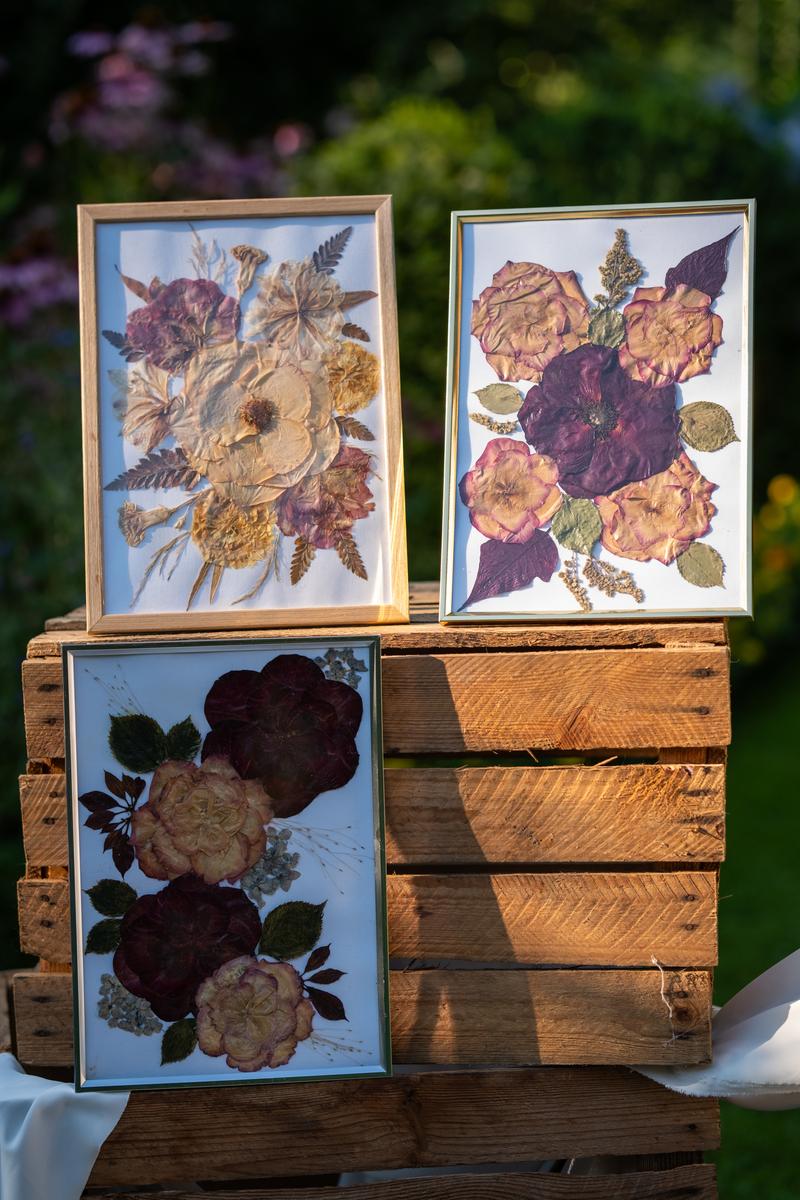 Trzy eleganckie obrazy z suszonych kwiatów w ramkach, ustawione na drewnianych skrzyniach w ogrodzie. Kolekcja Karokwiaty.