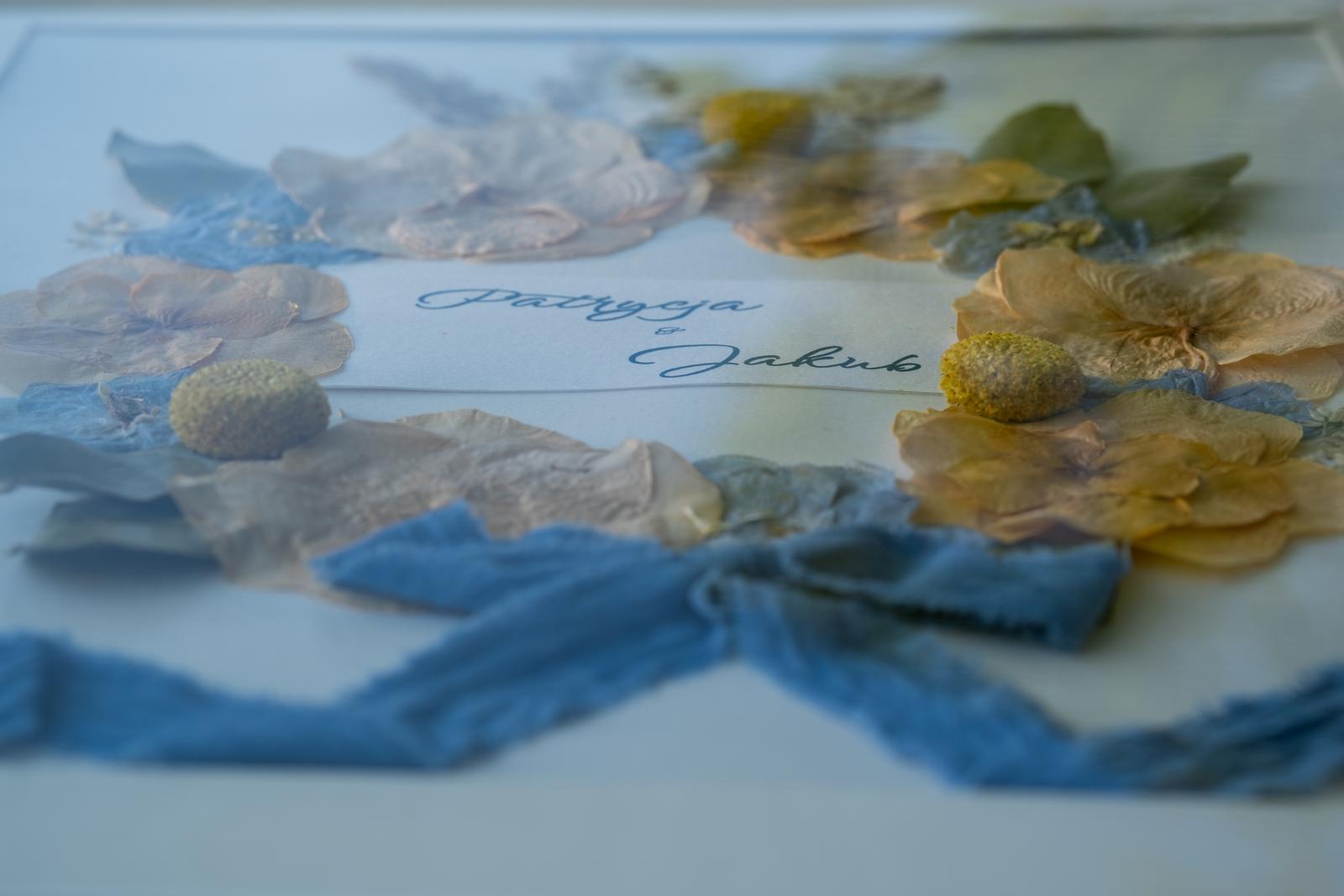 Zbliżenie na bukiet ślubny Patrycji i Jakuba w białej ramce. Pamiątka z suszonych płatków kwiatów i niebieską wstążką.
